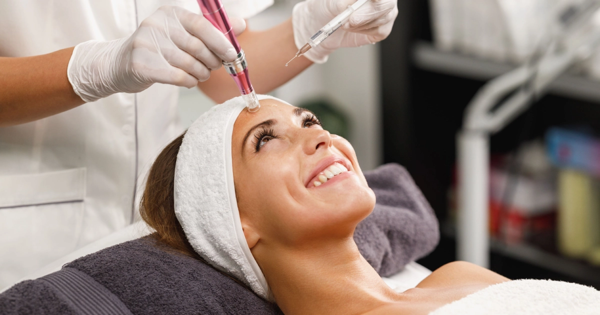 Smiling woman receives microneedling on her forehead from a practitioner in a softly lit clinic, hair and body covered with white towels in Hershey, PA