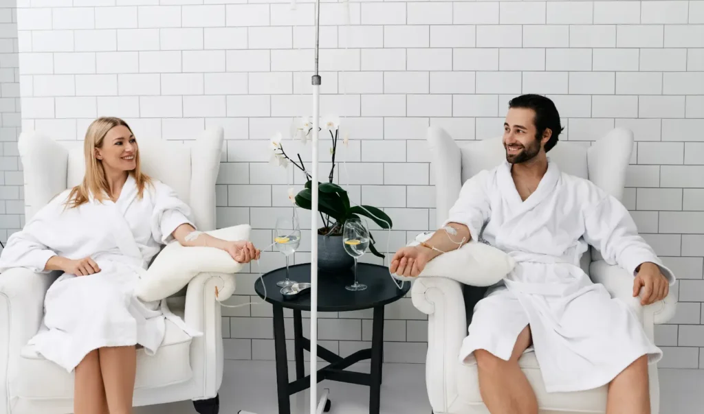 Two people in white robes receiving IV therapy while seated in a spa setting.