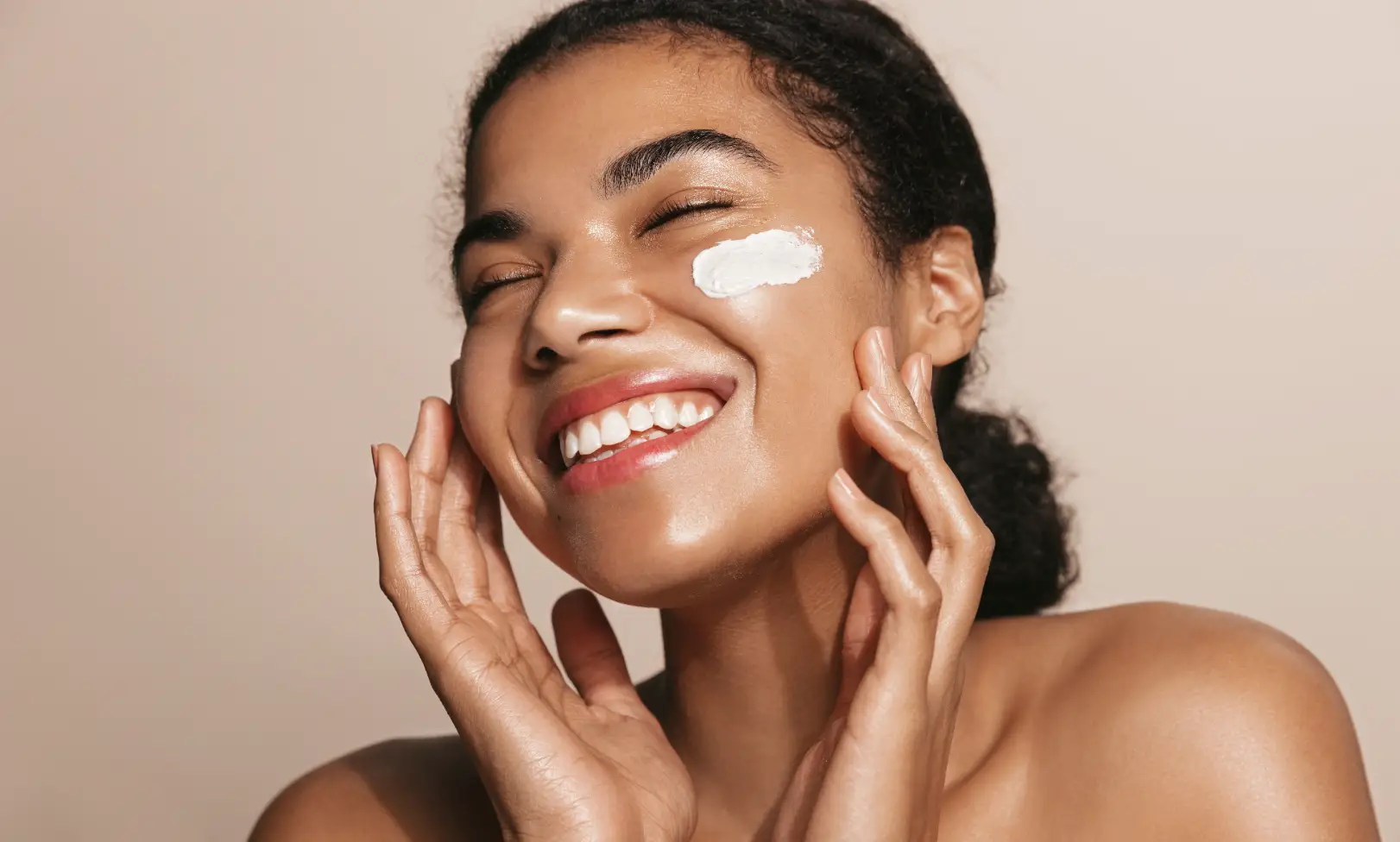 Person smiles while applying moisturizer to one cheek with fingertips against a neutral background.
