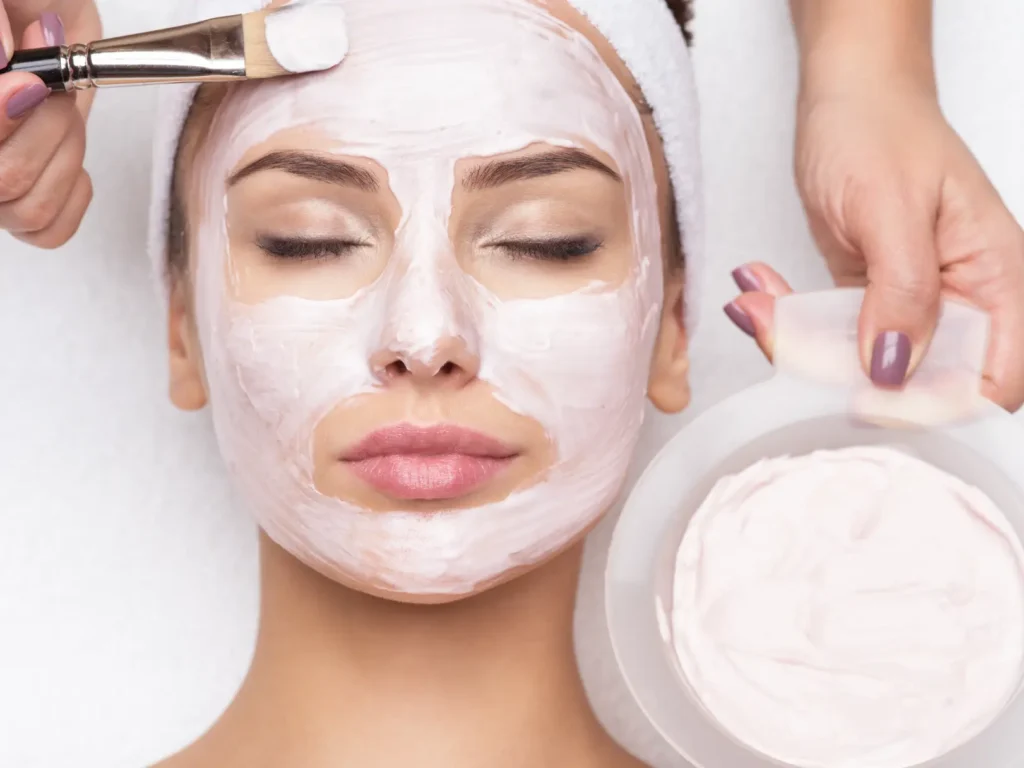 Facial mask is applied with a brush while a person rests with eyes closed on a treatment bed.