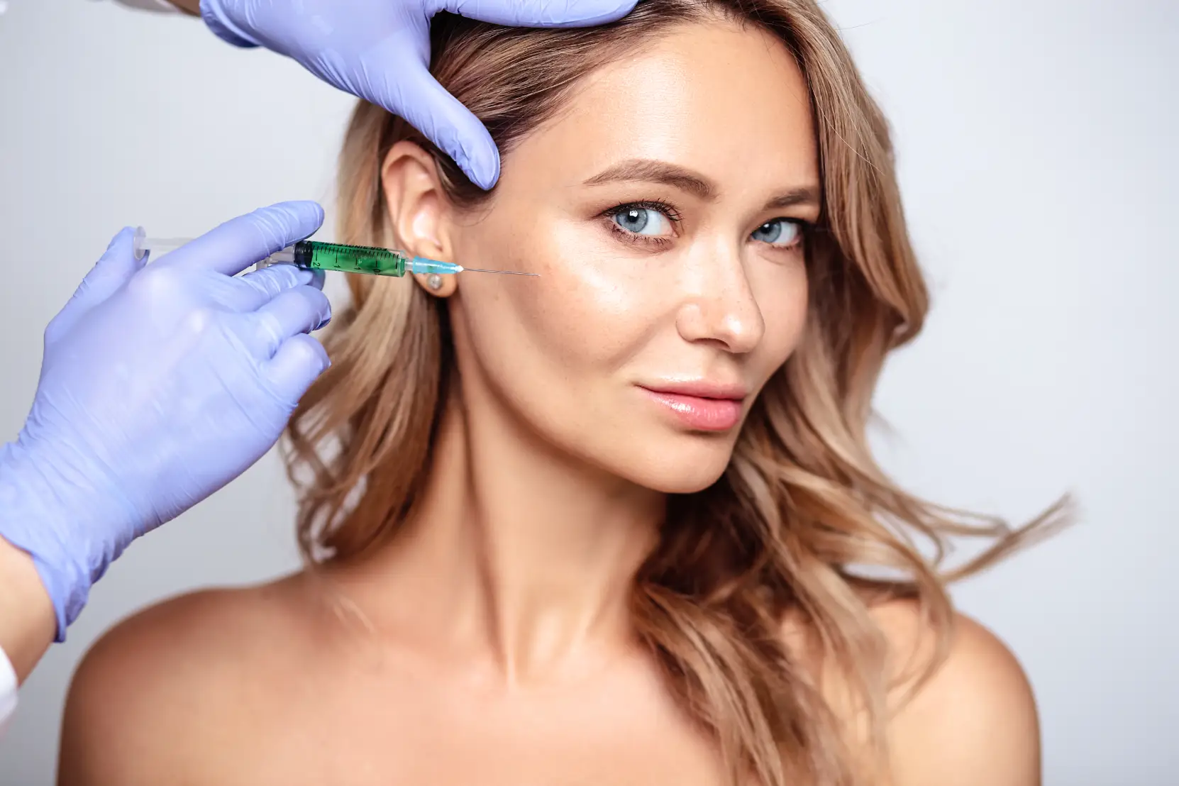 Practitioner positioning a syringe near a woman's cheek during a cosmetic injectable consultation.