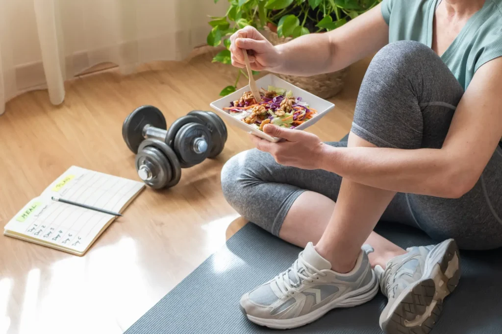 Woman in workout clothes eating a salad while seated on an exercise mat beside dumbbells and a meal planner.