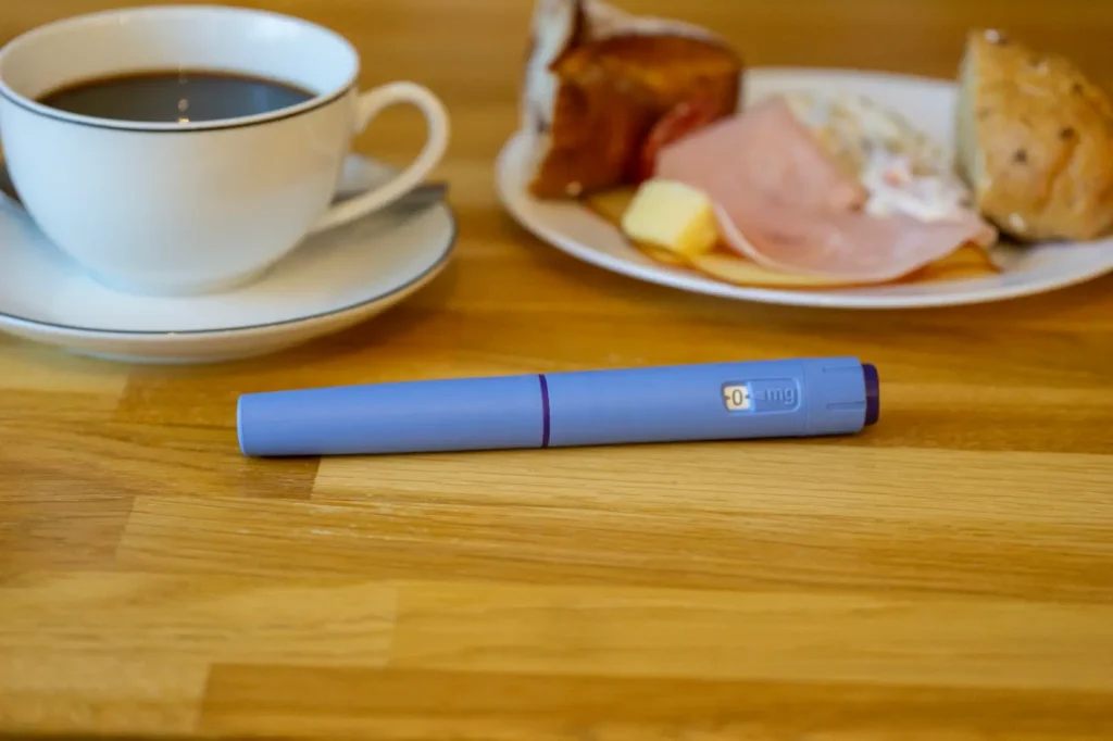 Blue injection pen on a wooden table with coffee, bread, ham, cheese, and other breakfast foods behind it.